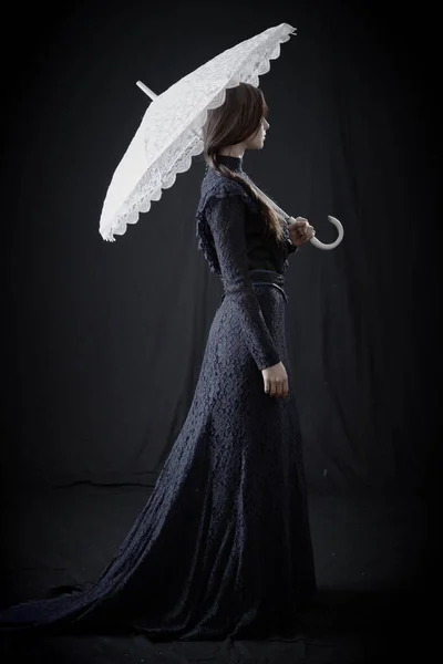 Eski elbiseli ve şapkalı bir kız koyu bir arka planda duruyor ve üzgün.