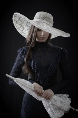 Eski elbiseli ve şapkalı bir kız koyu bir arka planda duruyor ve üzgün.