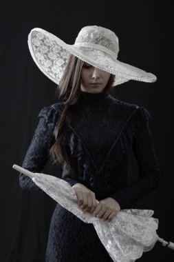 Eski elbiseli ve şapkalı bir kız koyu bir arka planda duruyor ve üzgün.