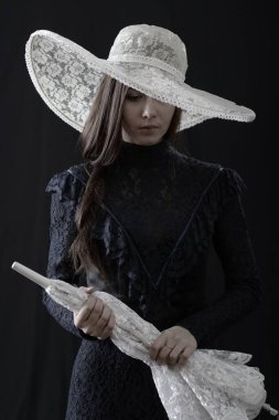 Eski elbiseli ve şapkalı bir kız koyu bir arka planda duruyor ve üzgün.