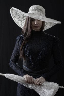 Eski elbiseli ve şapkalı bir kız koyu bir arka planda duruyor ve üzgün.