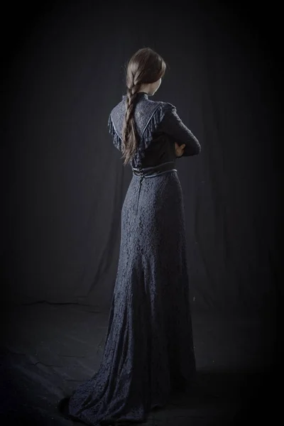 Eski elbiseli ve şapkalı bir kız koyu bir arka planda duruyor ve üzgün.