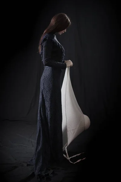 Eski elbiseli ve şapkalı bir kız koyu bir arka planda duruyor ve üzgün.