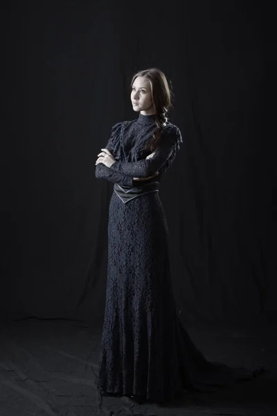 Eski elbiseli ve şapkalı bir kız koyu bir arka planda duruyor ve üzgün.