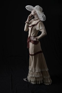 Eski elbiseli ve şapkalı bir kız koyu bir arka planda duruyor ve üzgün.