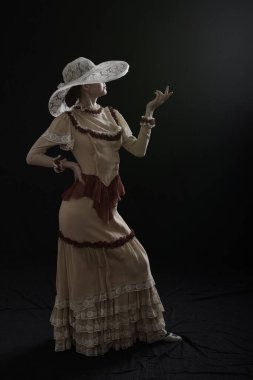 Eski elbiseli ve şapkalı bir kız koyu bir arka planda duruyor ve üzgün.