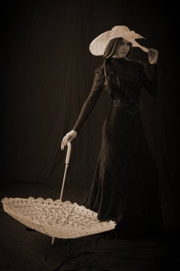 Eski elbiseli ve şapkalı bir kız koyu bir arka planda duruyor ve üzgün.