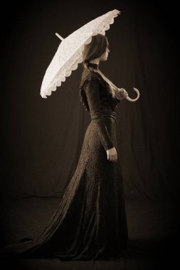 Eski elbiseli ve şapkalı bir kız koyu bir arka planda duruyor ve üzgün.