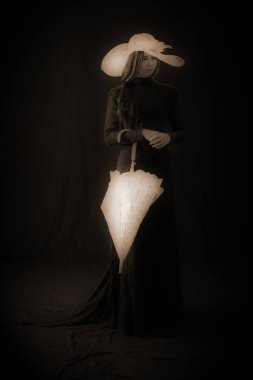 Eski elbiseli ve şapkalı bir kız koyu bir arka planda duruyor ve üzgün.