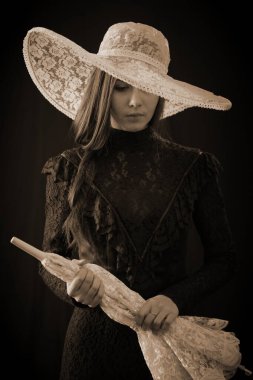 Eski elbiseli ve şapkalı bir kız koyu bir arka planda duruyor ve üzgün.