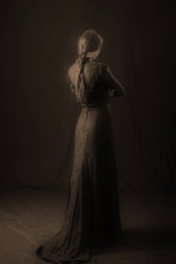 Eski elbiseli ve şapkalı bir kız koyu bir arka planda duruyor ve üzgün.