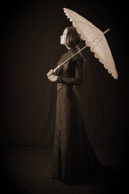 Eski elbiseli ve şapkalı bir kız koyu bir arka planda duruyor ve üzgün.