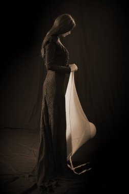 Eski elbiseli ve şapkalı bir kız koyu bir arka planda duruyor ve üzgün.