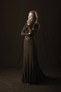 Eski elbiseli ve şapkalı bir kız koyu bir arka planda duruyor ve üzgün.