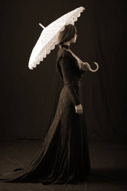 Eski elbiseli ve şapkalı bir kız koyu bir arka planda duruyor ve üzgün.