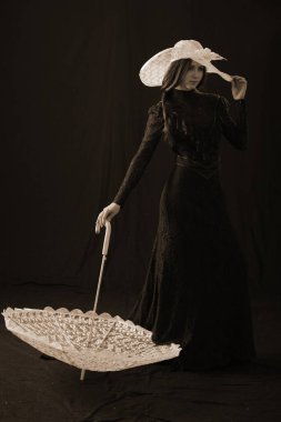 Eski elbiseli ve şapkalı bir kız koyu bir arka planda duruyor ve üzgün.