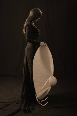 Eski elbiseli ve şapkalı bir kız koyu bir arka planda duruyor ve üzgün.