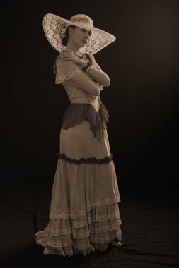 Eski elbiseli ve şapkalı bir kız koyu bir arka planda duruyor ve üzgün.