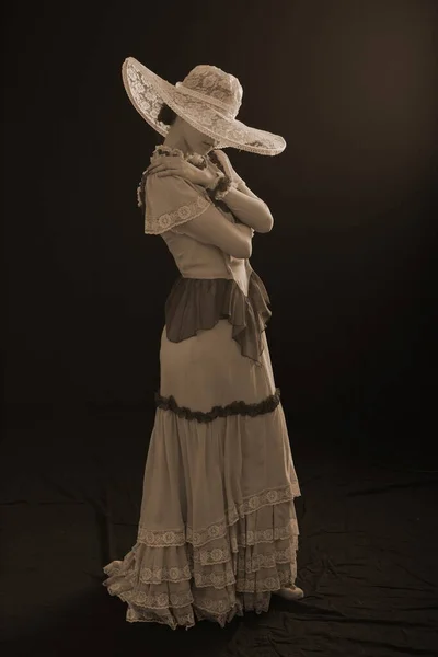 A girl in an old dress and hat stands on a dark background and is sad