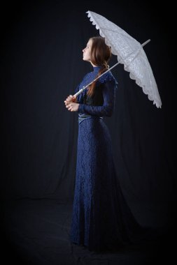 Eski elbiseli ve şapkalı bir kız koyu bir arka planda duruyor ve üzgün.