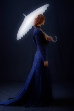 Eski elbiseli ve şapkalı bir kız koyu bir arka planda duruyor ve üzgün.