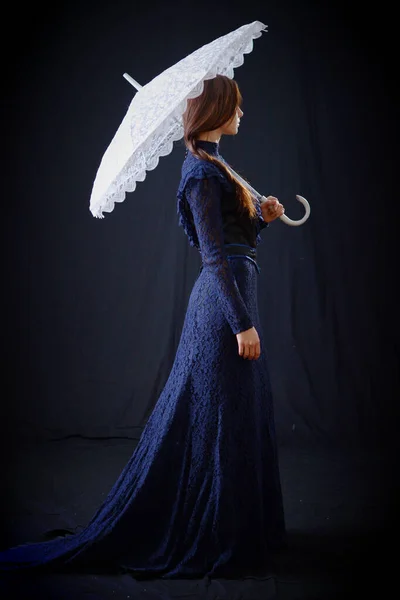 Eski elbiseli ve şapkalı bir kız koyu bir arka planda duruyor ve üzgün.