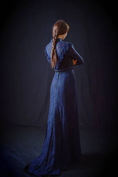 Eski elbiseli ve şapkalı bir kız koyu bir arka planda duruyor ve üzgün.