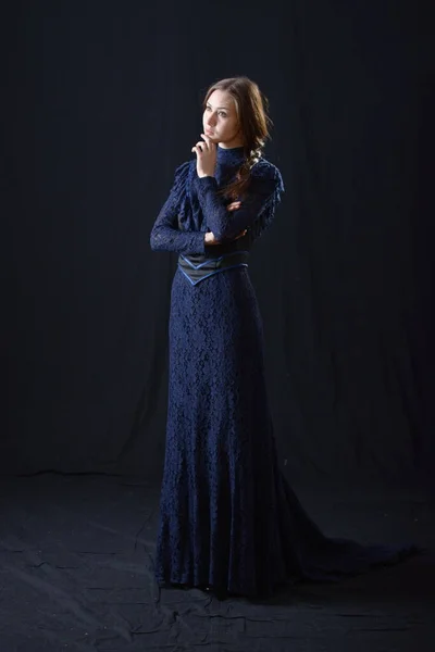 Eski elbiseli ve şapkalı bir kız koyu bir arka planda duruyor ve üzgün.