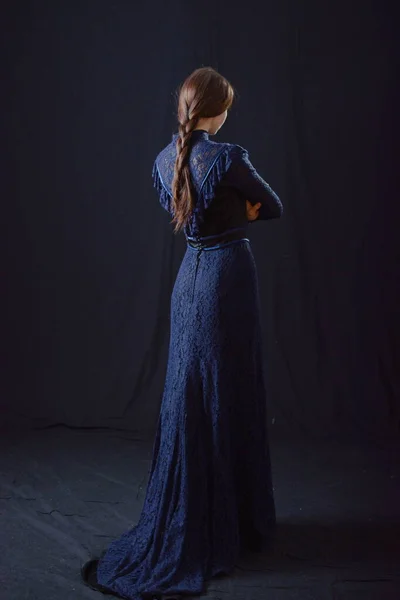 Eski elbiseli ve şapkalı bir kız koyu bir arka planda duruyor ve üzgün.