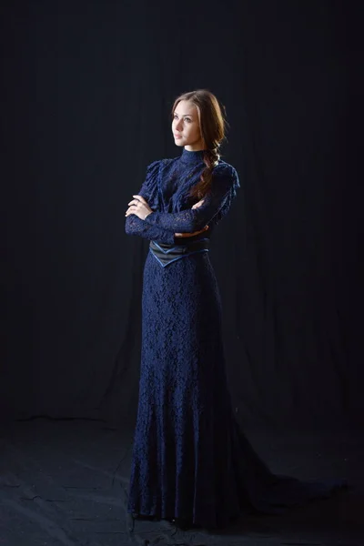 Eski elbiseli ve şapkalı bir kız koyu bir arka planda duruyor ve üzgün.