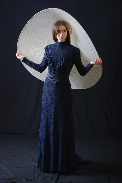 Eski elbiseli ve şapkalı bir kız koyu bir arka planda duruyor ve üzgün.