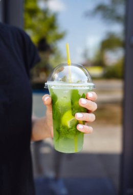 Şehir kafesinde elinde mojito olan plastik bardak tutan bir kadın. Buzlu meyve ve bitki süslemeli taze kokteyl içeceği. Seçici odak, bulanık arkaplan