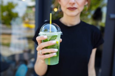 Şehir kafesinde elinde mojito olan plastik bardak tutan bir kadın. Buzlu meyve ve bitki süslemeli taze kokteyl içeceği. Seçici odak, bulanık arkaplan