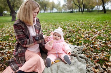 Güzel bir kız bebek ve annesi ekose üzerinde oturuyor. Aile açık havada. Güneşli bir günde, sonbahar parkında piknikte, kalın giysiler içinde sevimli küçük bir kız. Annesinin yanında güzel bir kız.