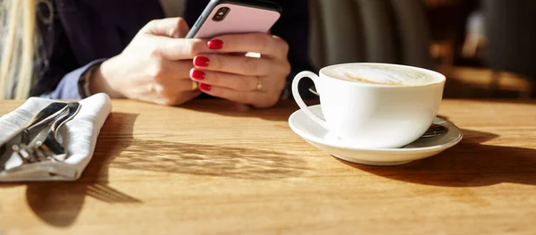 Bir kafede akıllı telefondan mesaj yazan bir kadın. Genç bir kadın kahve masasında cep telefonu kullanarak oturuyor. Kahve molası. Masanın üzerinde bir fincan kahve. Bulanık resim, seçici odak