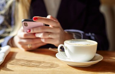 Bir kafede akıllı telefondan mesaj yazan bir kadın. Genç bir kadın kahve masasında cep telefonu kullanarak oturuyor. Kahve molası. Masanın üzerinde bir fincan kahve. Bulanık resim, seçici odak