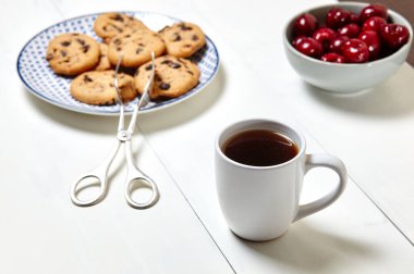 Beyaz fincanda kahve, geleneksel kurabiyeler ve beyaz ahşap arka planda seramik bir kasede tatlı kirazlar, yakın plan. Lezzetli kahvaltı, sabah rutini konsepti