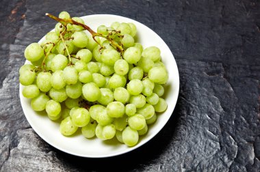 Tabakta su damlaları olan olgun yeşil üzüm dalı. Tahta arka planda sulu üzümler, yakın plan. Fotokopi alanı olan koyu mutfak masasında üzümler