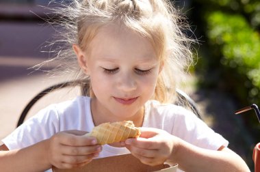 Küçük gülümseyen kız açık hava yazlık kafede kahvaltı yapıyor. Tatlı beyaz kız lezzetli waffle tüpü yiyor.