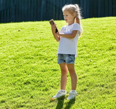 Yaz parkında çimlerin üzerinde duran ve elinde akıllı telefon tutan küçük bir kız. Çocukluk, eğlence ve insan konsepti. Mutlu çocuk istirahati ve iyi vakit geçirin.