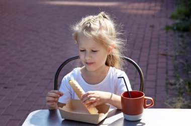 Küçük gülümseyen kız açık hava yazlık kafede kahvaltı yapıyor. Tatlı beyaz kız lezzetli waffle tüpleri yiyor ve kakao içiyor.