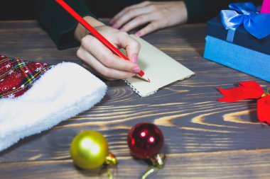 Kadın Noel kartpostalları yazıyor. Bayramlarda selamlar. Yeni yılınız kutlu olsun. Arkadaşlarını ve aileni tebrik ederim. Tahta masadaki dekorasyonlar. Keyfim yerinde. Boşluğu kopyala.