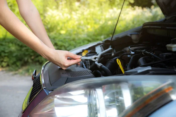 Woman is repairing broken car. Accident situation while travelling. Opened bonnet of vehicle. Girl is holding instruments for fixing. Doesn;t know what to do.