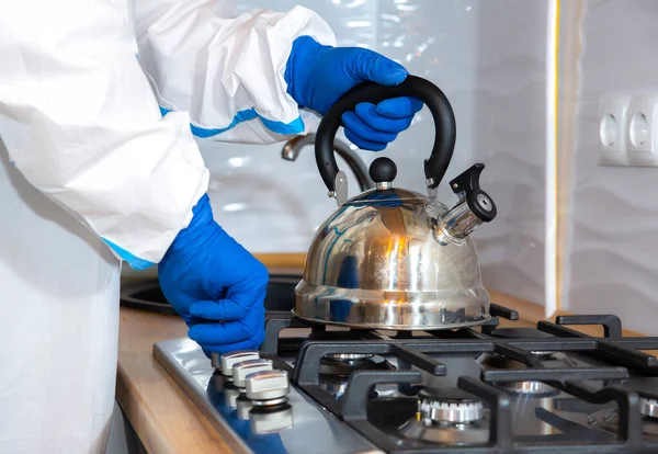 Doctor wearing medical protective suit, goggle, mask, and gloves preparing tea on the kitchen. Teapot on the gas stove. Protection mers by virus epidemic. Coronavirus (COVID-19). Healthcare concept.