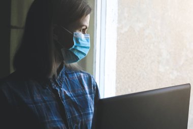 Tıbbi maskeli kadın evde çalışıyor. Karantina zamanında online serbest çalışmalar. Dizüstü bilgisayar pencerenin yanında. Coronavirüs pandemisi. Ekonomik kriz alternatifleri.