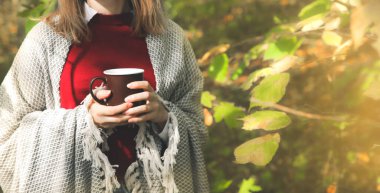 Güz ormanında kahve içen güzel bir kız. Sıcak bir bardak. Keyifli bir ortam. Kırmızı ve sarı yapraklar. Güzel doğa. Kadınların omuzlarında şık bir battaniye.