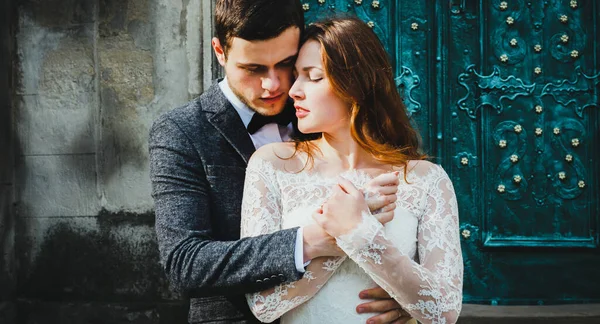 Wedding couple hugs near the vintage green door. Stone walls in ancient town background. Rustic bride with hair down in lace dress and groom in grey suit and bow tie. Tender embrace. Romantic love.