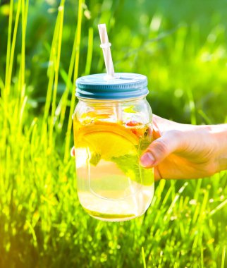 Girl holding fresh lemonade in jars with straws. Hipster summer drinks. Eco-friendly in the nature. Lemons, oranges and berries with mint in the glass. Green high grass outdoors.