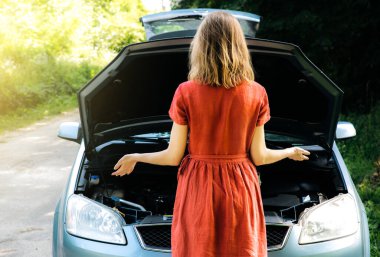 Kadın, doğadaki bozuk arabanın yanında duruyor. Seyahat sırasında kaza durumu. Elbiseli kız ne yapacağını bilmiyor. Araç kaputunu aç.