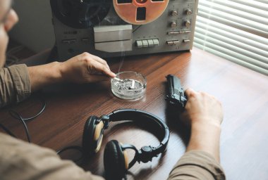 Özel ajan bobin kayıt cihazını dinliyor. Memur sigara içiyor. KGB konuşmaları gözetliyor. Kül tablasının yanında sigaralı bir el. Masada silah var..
